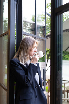 Thoughtful relaxed businesswoman restaurant administrator enjoying aromatic coffee on lunch break during workday. Confident female boss sipping, standing at entrance of terrace.