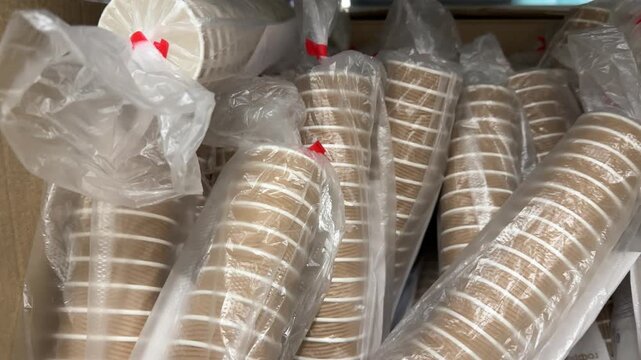 Stacks of disposable plastic drinking cups are packed in transparent bags and cardboard boxes, neatly arranged for sale in a large retail store for parties, events and everyday beverage service.