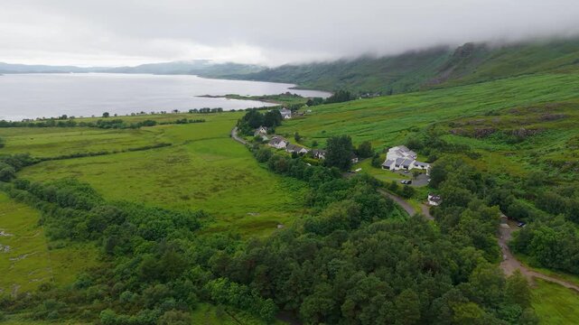 Drone flight over croft houses, green fields and a winding road beside Loch Torridon with surrounding Highland mountains partly hidden by low cloud