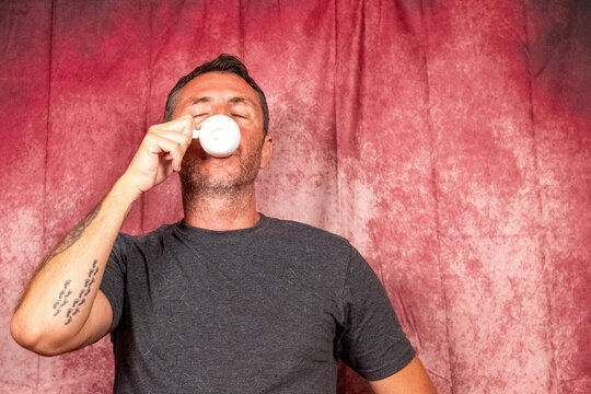 A man drinks an Italian coffee from a small white porcelain cup. The man is forty-two years old, has short hair and a short beard and wears a dark T-shirt