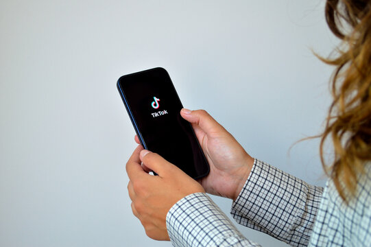 Young woman using a black smartphone displaying the TikTok social media app splash screen, wearing a checkered shirt against a clean light gray background
