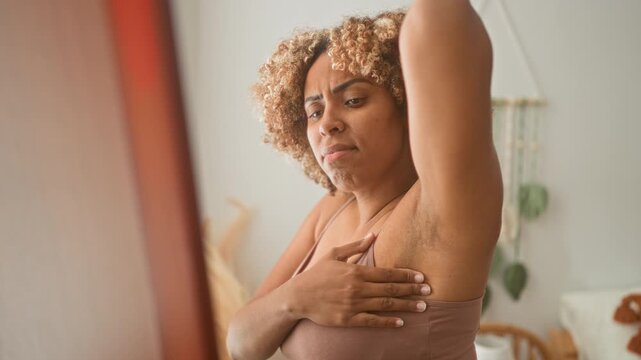 Woman checks armpit in bedroom mirror, raising arm and touching underarm skin while inspecting for lumps and irritation; self care concern.