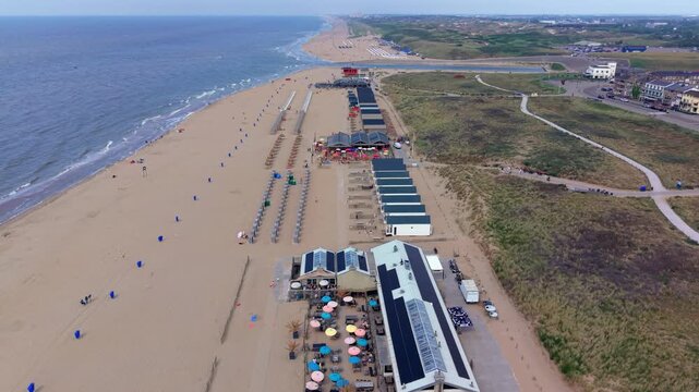 Aerial view video clip of Katwijk aan Zee coastline in Netherlands, featuring beach pavilions, solar panels, beach cabins, sand dunes, and North Sea waves on a sunny day