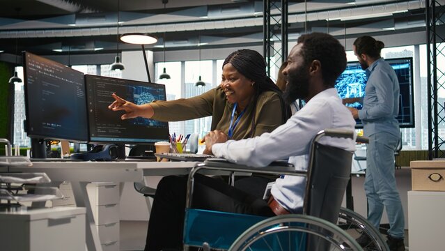 Startup office programmer in a wheelchair talks with a coworker while developing artificial intelligence models, running analytics simulations, and testing software for AI forecasting.