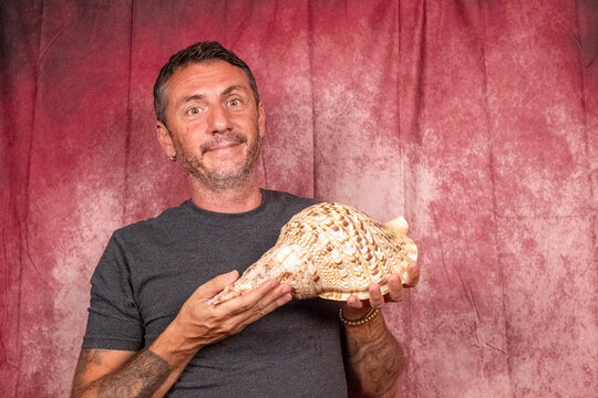 A man shows off a large sea shell. The man is forty-two years old and wears a dark T-shirt