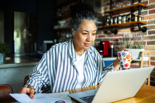 Mature woman working from home kitchen with laptop and coffee