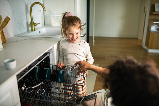 Smiling girl helping parent unload dishwasher in modern home kitchen