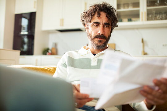 Bearded man reviewing household bills with laptop in home kitchen