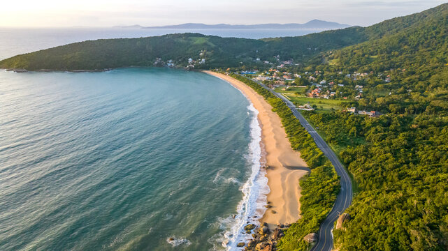 Aerial view of Praia das Laranjeiras featuring a sandy beach, turquoise ocean waves, a coastal road, and lush green hills in Balneario Camboriu, Santa Catarina, Brazil.