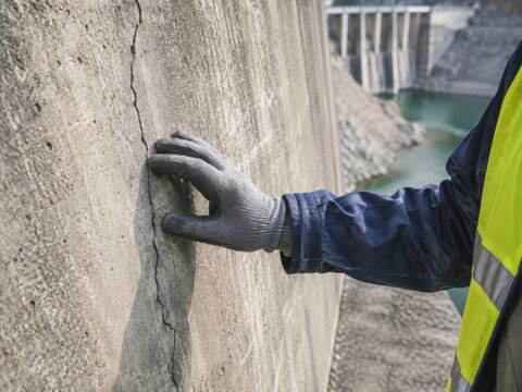 A close-up view of a worker in protective gear inspecting a vertical crack in a large concrete wall.