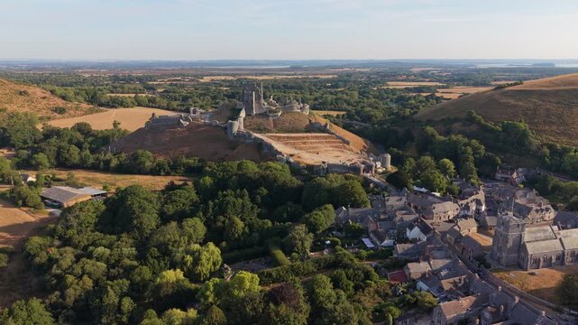 Aerial drone footage of the iconic Corfe Castle in Dorset, England, showcasing the medieval castle ruins, historic village, and rolling Purbeck countryside on a hot summer morning
