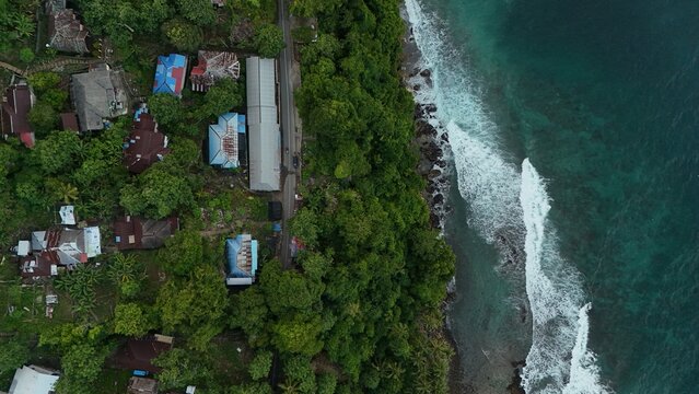 Aerial view of Tengah-Tengah village with coastal houses, lush tropical trees, a winding road, and turquoise waters of the Banda Sea hitting the rocky shore in Salahutu, Maluku, Indonesia.