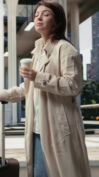 Vertical frame: Female passenger walks to the coach departure area with a coffee cup and trolley; the woman carries takeaway coffee and luggage through the terminal before an intercity bus journey.
