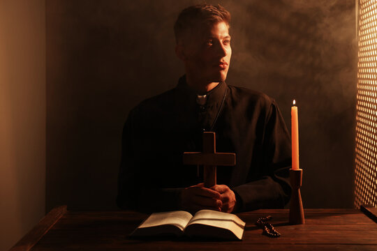 Priest holding cross at wooden table with Holy Bible, rosary and candle in confessional booth. Space for text