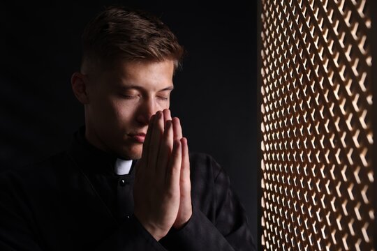 Priest with clasped hands praying near partition in confessional booth