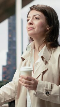 Vertical frame: Close-up: A female passenger walking to the coach departure area with a coffee cup and trolley, carrying takeaway coffee and luggage through the terminal for an intercity bus journey.