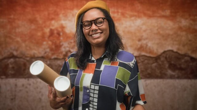 Young african american woman holding rolled blueprints and smiling, hand near face and wearing beret against a textured building wall; creative confidence.