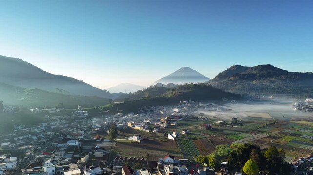 Aerial view of Dieng Plateau featuring a village nestled among misty mountains and agricultural fields under a clear morning sky in Indonesia.