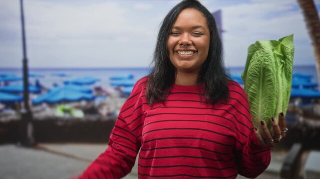Young woman in red striped sweater holds romaine lettuce in right hand and shrugs left palm up at crowded beach promenade, smiling with septum ring; joy wellness.
