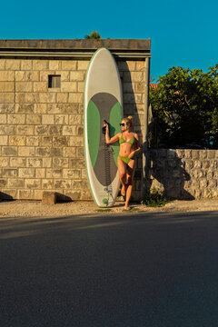 A blonde woman posing in olive green bikini and sunglasses stands in profile next to a large green and white stand-up paddleboard