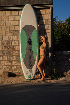 A blonde woman wearing an olive green bikini and sunglasses stands in profile next to a large green and white stand-up paddleboard