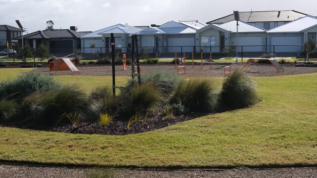 Off-leash dog agility park featuring ramps, hurdles, and weave poles set inside a fenced enclosure in Tarneit, Melbourne, Australia. Suburban houses line the background. Community recreation facility