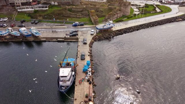Aerial view of a small working fishing boat moored at a harbor pier, surrounded by seagulls, fishing equipment, parked vehicles and rows of colorful boats along the waterfront.