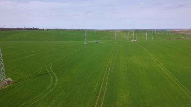 Aerial approach over a broad green crop field crossed by electricity pylons, with a distant agricultural sprayer traveling along parallel crop tracks.