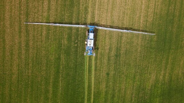 Top down aerial tracking view of a blue crop sprayer with extended booms moving along parallel tracks through a green field with lighter planted strips.