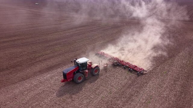 Aerial view of tractors performing field preparation and precision planting across a large cultivated agricultural field, using wide implements that create parallel rows and raise dust behind the mach