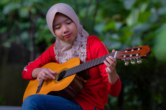 Teenage Indonesian girl wearing a headscarf sitting outdoors playing the guitar