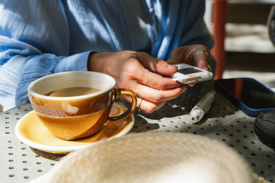 Close-up of a diabetic woman sitting at an outdoor cafe table with a cup of white coffee checking her glaucometer readings