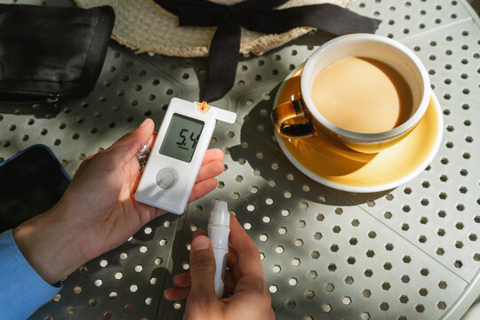 Personal perspective view of a diabetic woman sitting at an outdoor cafe table with a cup of white coffee checking her glaucometer readings