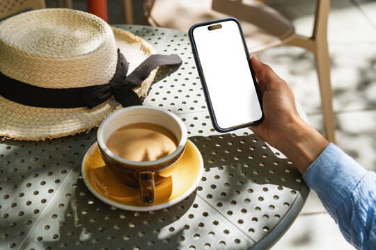 Personal perspective view of a woman sitting at an outdoor cafe holding a mobile phone with a blank screen and drinking a cup of white coffee on a summer day