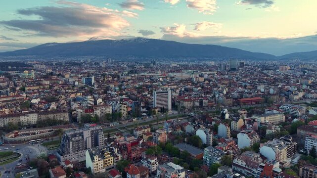 Aerial view of Sofia, Bulgaria, with dense residential neighborhoods, modern apartment blocks, tree-lined streets and parks spreading toward the foothills of Vitosha Mountain under soft evening light.