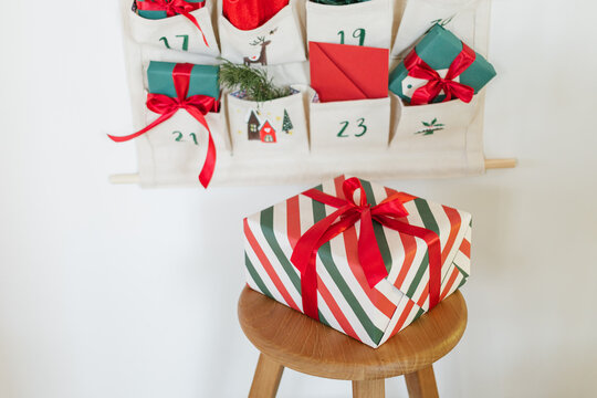 Close-up of part of a Traditional Christmas advent calendar with assorted wrapped gifts hanging on a white wall next to a wooden stool with a larger wrapped Christmas gift