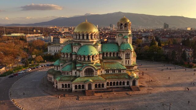 Aerial golden hour view of Alexander Nevsky Cathedral in Sofia, Bulgaria, highlighting its monumental golden dome, Orthodox cross, green-roofed towers and surrounding city center architecture.