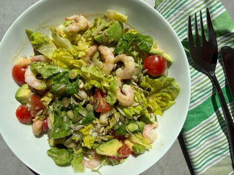Overhead view of a bowl of salad with prawn, avocado, pumpkin and sunflower seeds on a table with cutlery and a napkin