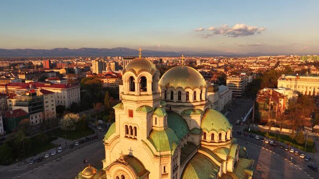 Aerial golden hour view of Alexander Nevsky Cathedral in Sofia, Bulgaria, highlighting its monumental golden dome, Orthodox cross, green-roofed towers and surrounding city center architecture.