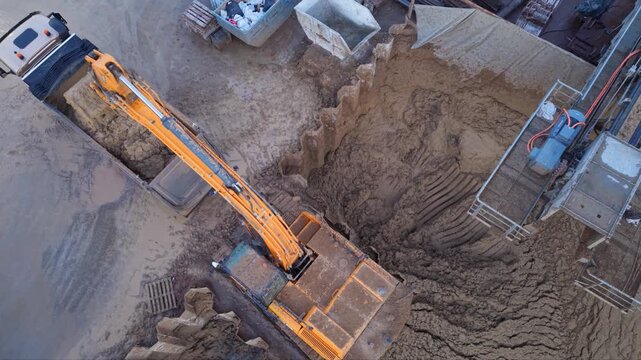Close aerial orbit above an excavator transferring soil into a dump truck, with its long boom reaching across a stockpile beside a deep braced construction site.