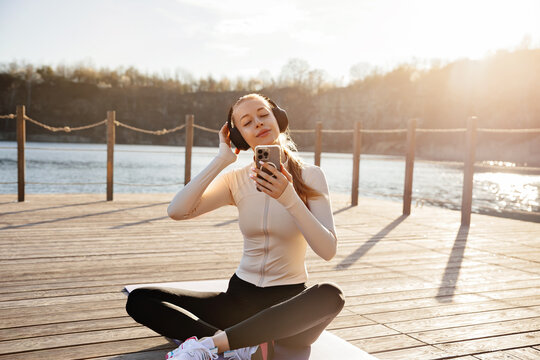 Pretty woman enjoying music after outdoor fitness session. Portrait of female model relaxing on yoga mat. Wellness concept.