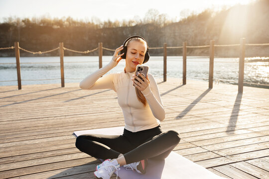 Beauty woman relaxing after yoga while listening to music. Young woman sitting on fitness mat with smartphone outdoors. Active lifestyle concept.