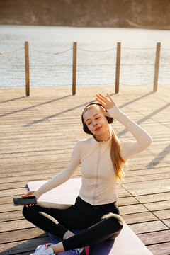 Beautiful woman listening to music after yoga workout. Beauty woman relaxing on fitness mat with headphones outdoors. Wellness concept.