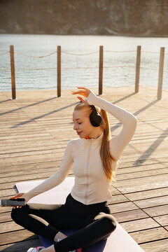 Young woman relaxing on yoga mat with headphones. Pretty woman checking smartphone after workout outdoors. Fitness lifestyle concept.