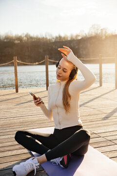 Beautiful woman enjoying music while sitting on yoga mat. Attractive woman using smartphone after outdoor exercise. Active lifestyle concept.