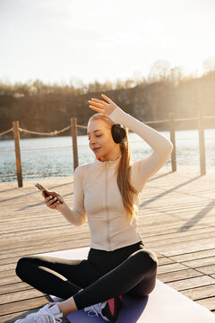 Beauty woman smiling with headphones during yoga break. Young woman holding smartphone beside peaceful waterfront. Wellness concept.