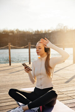 Attractive woman listening to music on yoga mat. Beauty woman checking smartphone with relaxed smile outdoors. Healthy lifestyle concept.