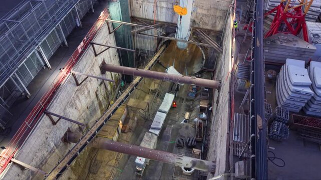 Aerial view inside a deep industrial construction shaft where workers install large ventilation duct sections beneath heavy steel bracing, surrounded by concrete walls, lifting cables and construction