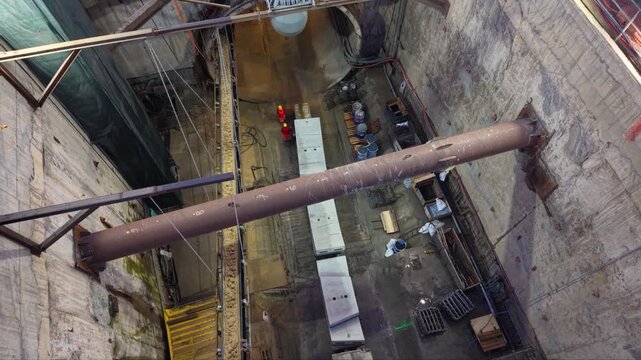 Aerial view inside a deep industrial construction shaft where workers install large ventilation duct sections beneath heavy steel bracing, surrounded by concrete walls, lifting cables and construction