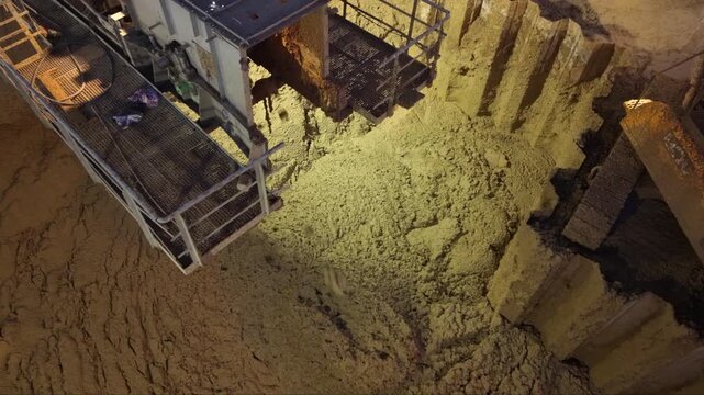 Overhead night view of an excavator reaching into a floodlit soil pile beside a machinery platform, with steel retaining piles bordering the dark construction site.